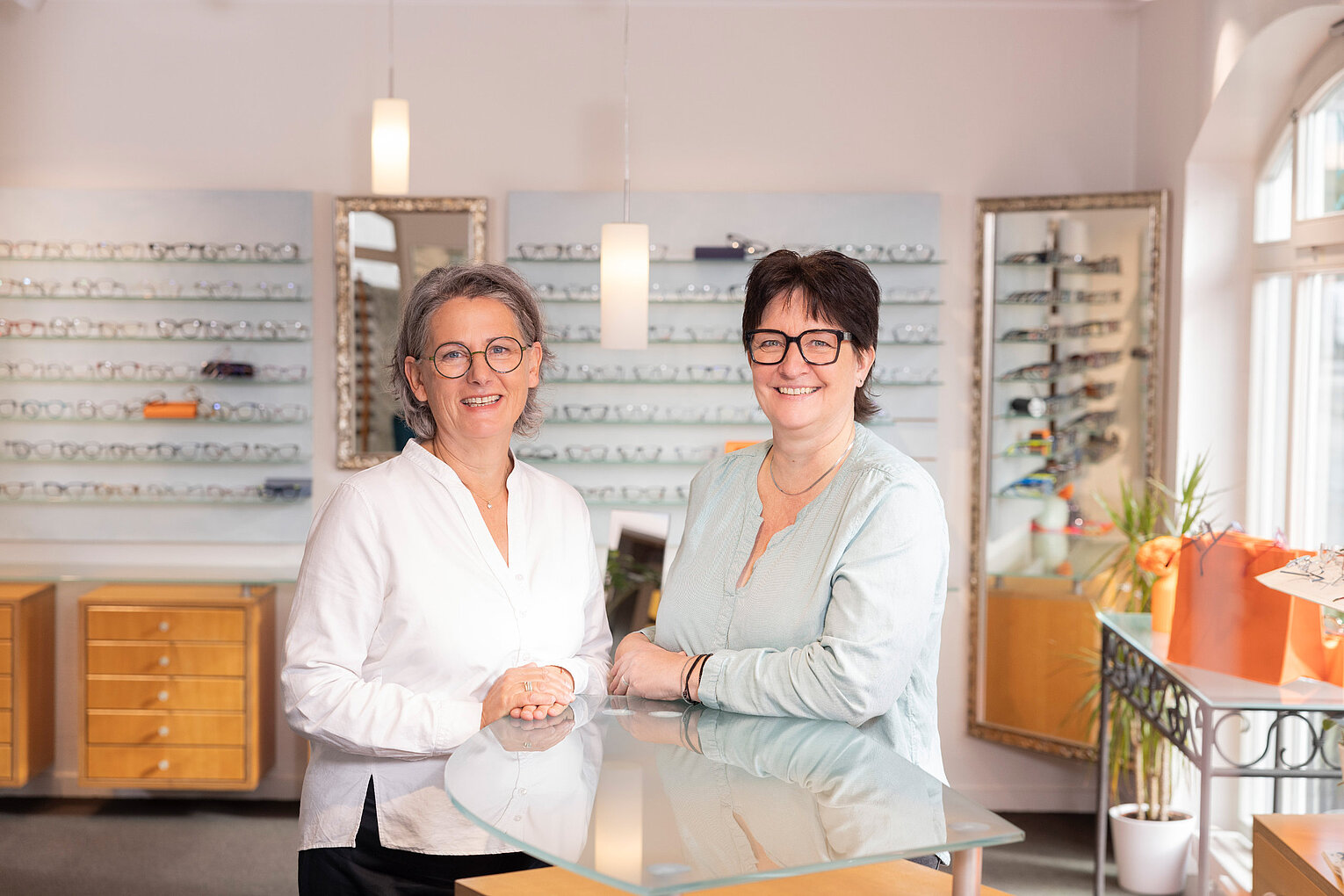 Kompetente Beratung im stilvollen Optikgeschäft Zwei Mitarbeiterinnen stehen lächelnd hinter einem Glastisch im stilvoll eingerichteten Optikgeschäft mit Brillenauslage.